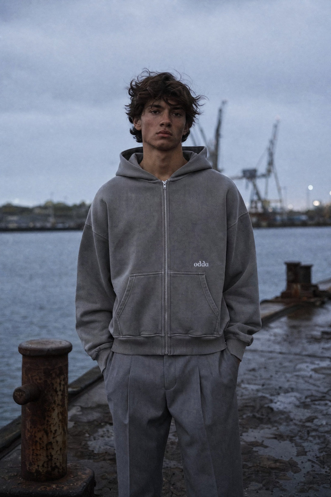 Young man in a grey Odda zip hoodie by the harbour at dusk